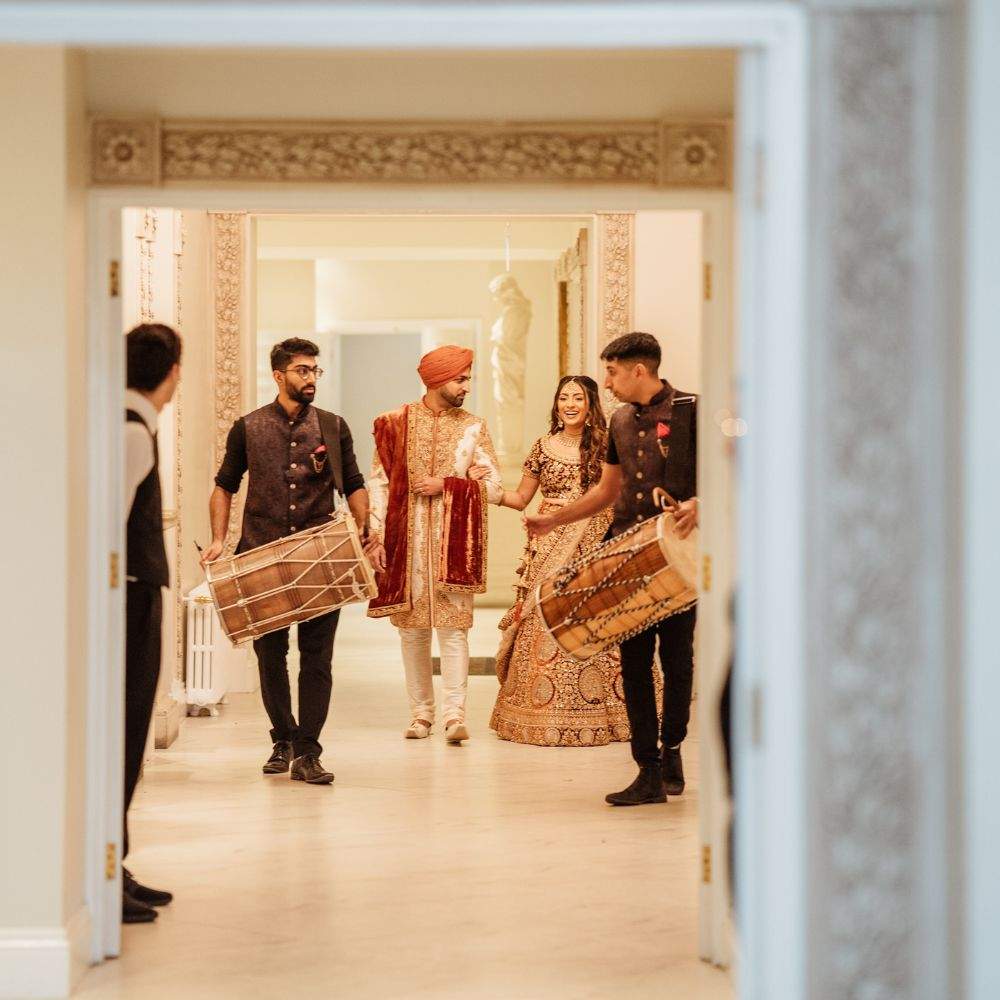 A Sikh Bride & Groom dressed in colourful cultural attire being drummed into their Sikh wedding reception by Dhol drummers at Froyle Park.