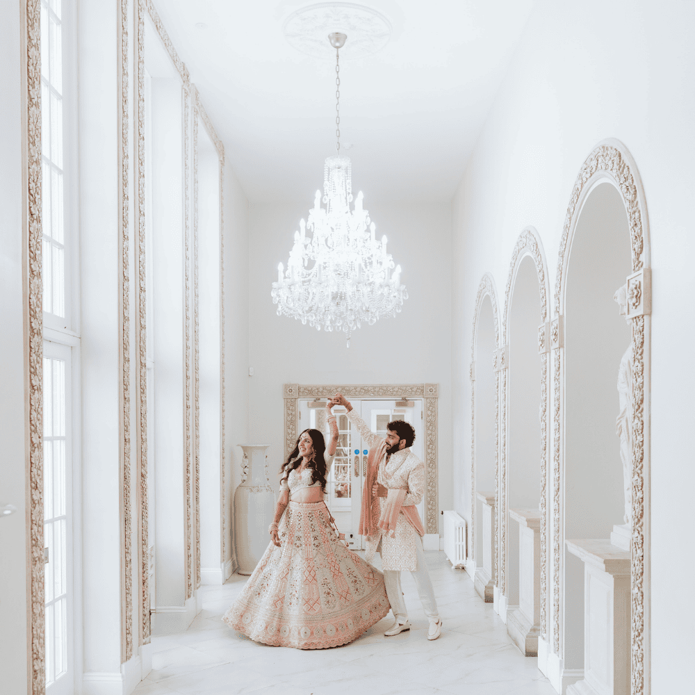 An Asian bride and groom, dressed ceremonial outfits, twirl in the light-filled Grand Ballroom walkway at Froyle Park