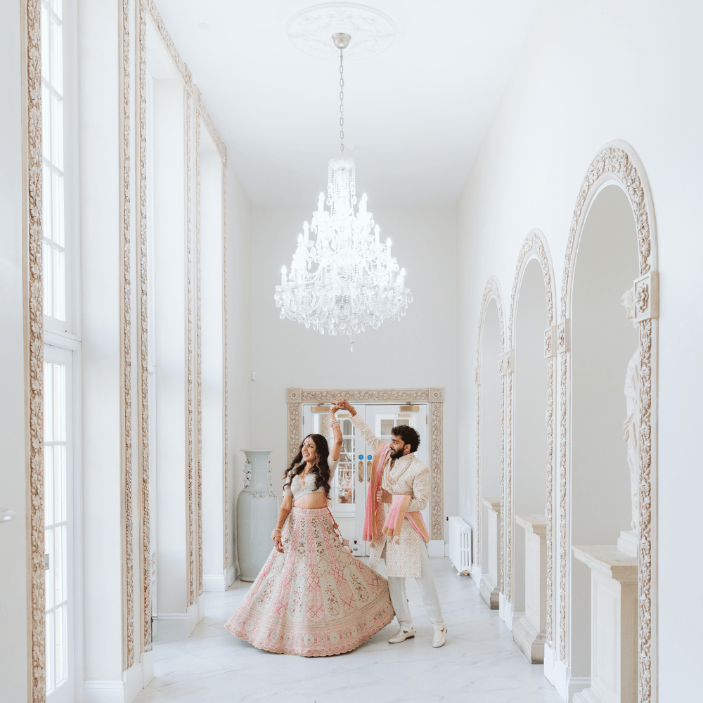 An Asian bride and groom, dressed ceremonial outfits, twirl in the light-filled Grand Ballroom walkway at Froyle Park