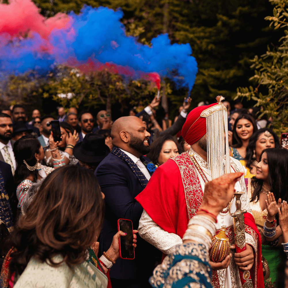 An Asian groom arrives at Froyle Park, the UK's finest South Asian Wedding Venue, during his Baraat grand entrance with colourful smoke flares and guests celebrating his arrival