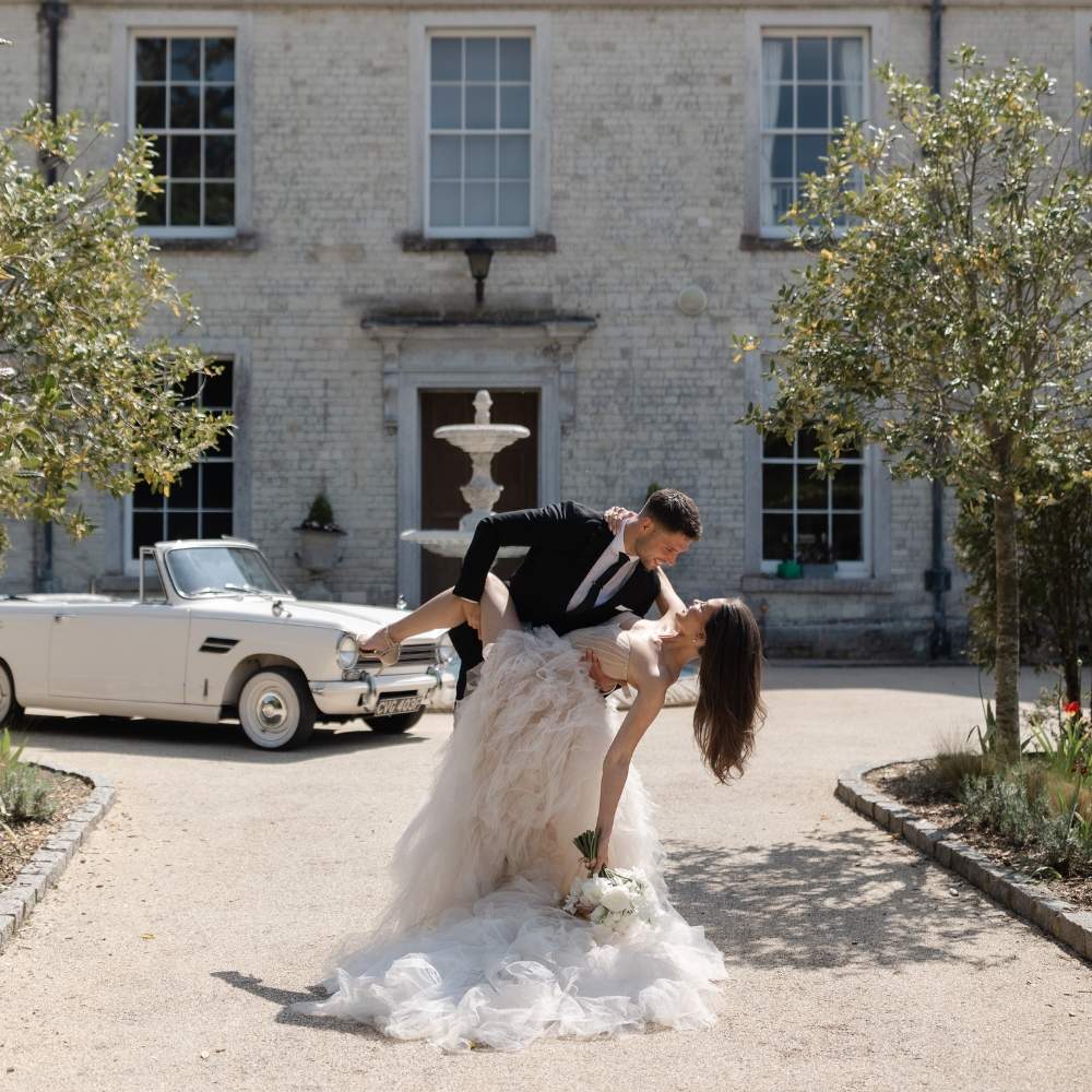An elegant bride and groom pose in front of the manor house at Froyle Park. A vintage car can be seen in the background