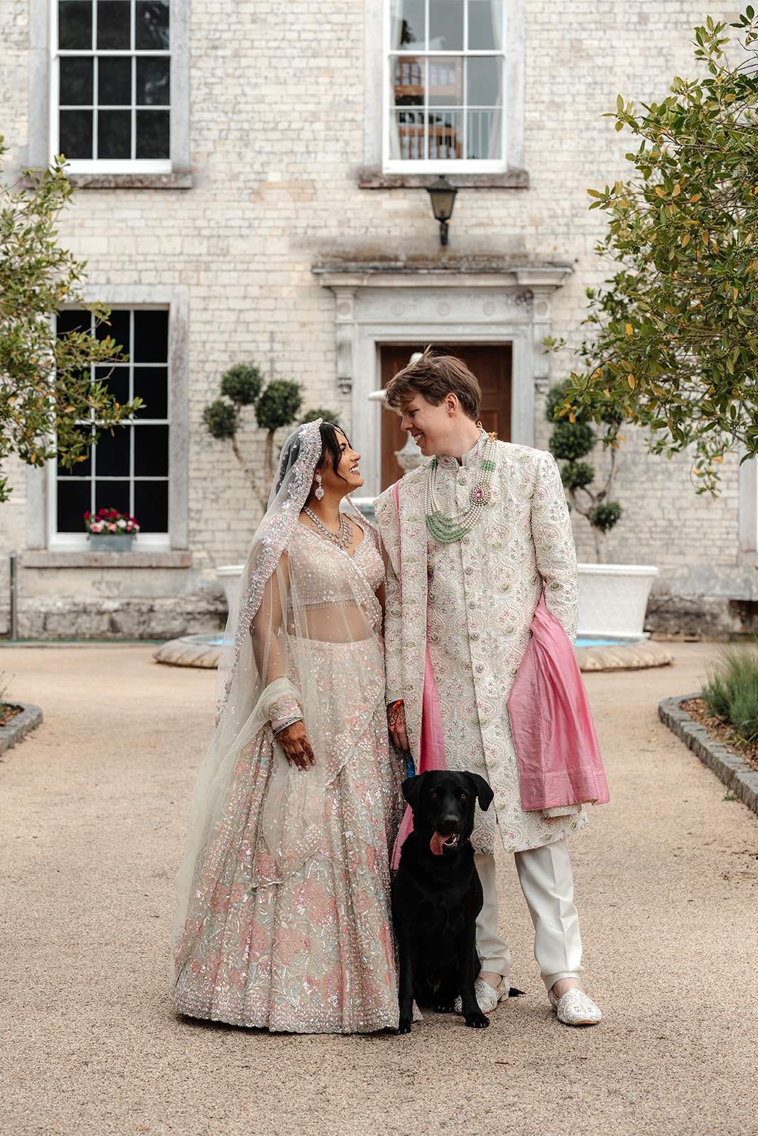 A bride and groom in traditional Hindu wedding outfits stand in Front of Froyle Park's Manor house with their pet Labrador