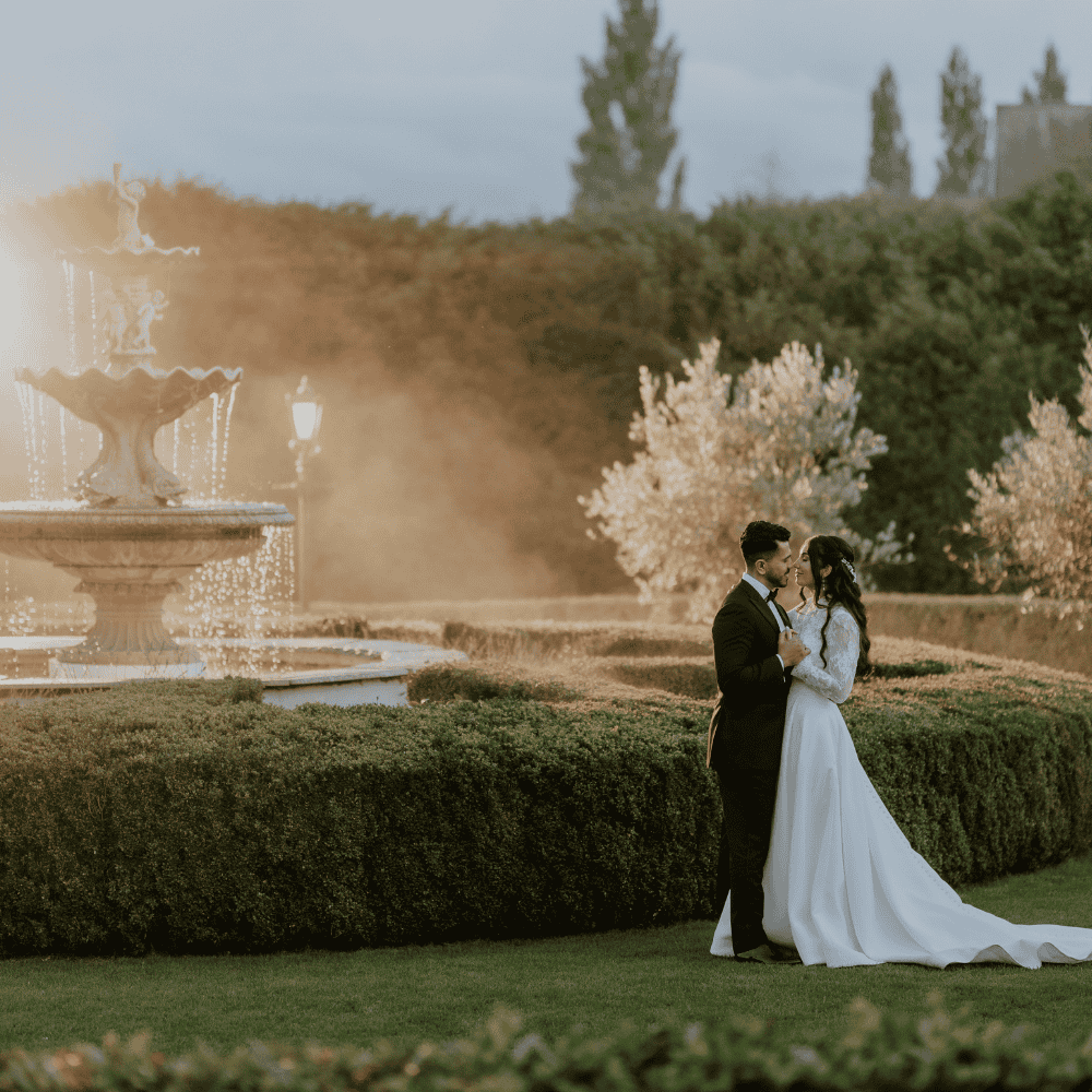 Following an outfit change into black tie, an Asian Bride & Groom pose for photos in the grounds of Froyle Park beside the ornate water fountain.